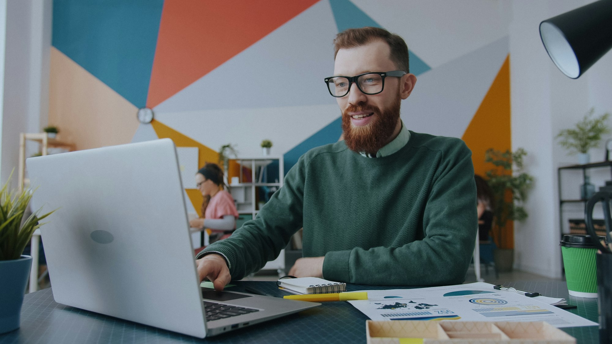 A smiling man with glasses working on a laptop in a colourful office